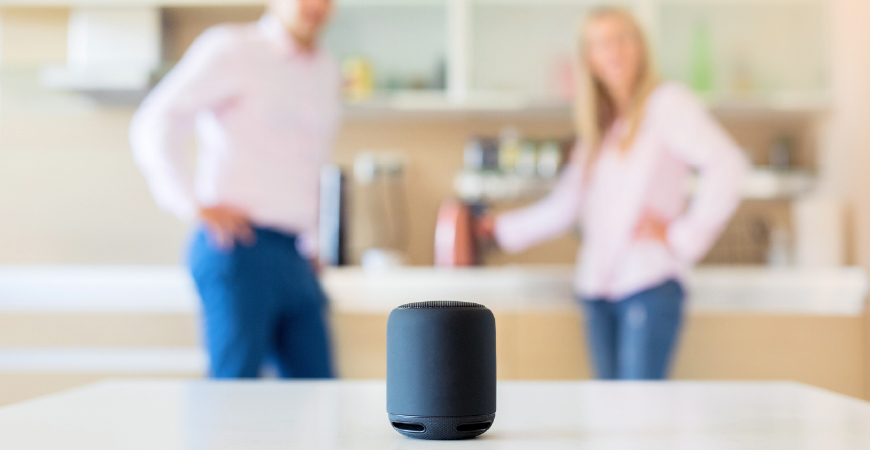Couple using smart speaker at home blurred, with focus on speaker in the foreground.